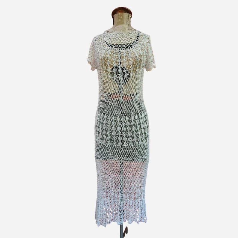 K&ouml;nnte beinhalten: Wei&szlig;es H&auml;kelkleid mit offenem Gewebe, kurzen &Auml;rmeln und Midi-L&auml;nge. Das Kleid wird auf einer Schaufensterpuppe pr&auml;sentiert, die das komplizierte Muster und die zarte Textur zeigt. Das Kleid hat einen gewellten Saum.