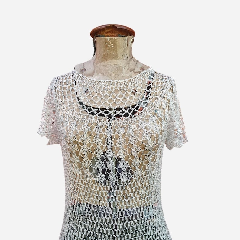 K&ouml;nnte beinhalten: Ein wei&szlig;es, kurz&auml;rmeliges H&auml;keltop mit rundem Ausschnitt. Das offene Design des Tops l&auml;sst einen Blick auf die darunterliegende Unterw&auml;sche zu. Das Top wird auf einer Vintage-Mannequin pr&auml;sentiert.