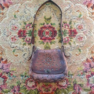 May include: A brown leather shoulder bag featuring a flap with intricate laser-cut floral patterns and a central metal turn-lock closure. The bag has a single leather strap and visible stitching along the edges of the rounded base.