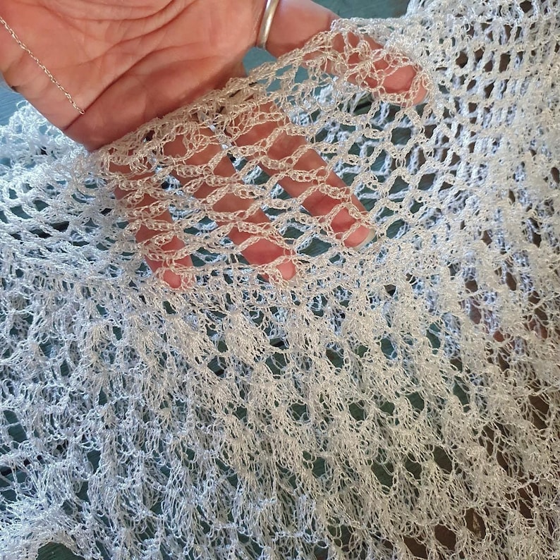 K&ouml;nnte beinhalten: Nahaufnahme eines zarten, offen gewebten, wei&szlig;en H&auml;kelstoffs. Das komplizierte Muster erzeugt einen Spitzeneffekt mit kleinen Quasten am Rand. Der Stoff wirkt weich und leicht, geeignet f&uuml;r einen Schal oder Schal.