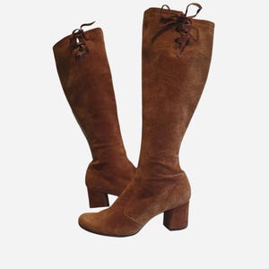 Könnte beinhalten: Ein Paar kniehohe braune Wildlederstiefel mit Blockabsatz und Schnürung am oberen Schaftrand. Die Stiefel haben eine abgerundete Zehenpartie und eine strukturierte Oberfläche, wobei passende braune Schnürsenkel hinten an der Öffnung gebunden sind.