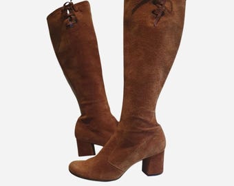 Puede incluir: Un par de botas altas de ante marrón hasta la rodilla con tacón de bloque y detalle de cordones en la parte superior. Las botas tienen una puntera redondeada y un acabado texturizado, con cordones marrones a juego atados en la parte trasera.