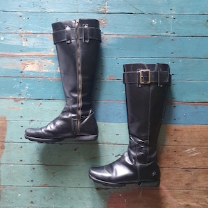 May include: Two black leather knee-high boots. One boot has a full-length zipper and a buckle strap at the top. The other boot has a buckle strap at the top. The boots are displayed against a weathered, multi-colored wooden background.