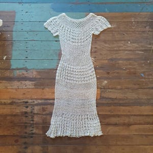 May include: A light beige, handmade crochet dress with short sleeves and a flared skirt. The dress features a delicate open weave pattern throughout, creating a textured appearance. The dress is displayed on a wooden surface.