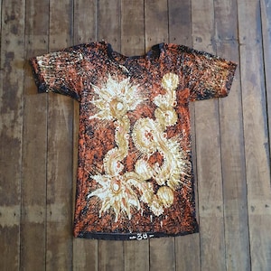 Könnte beinhalten: Ein kurzärmeliges T-Shirt mit Rundhalsausschnitt und einzigartigem Design. Das Shirt hat einen braunen und schwarzen gesprenkelten Hintergrund mit einem großen, verzierten, gold-weißen Blumenmuster. Die Nummer "38-" ist unten aufgedruckt.