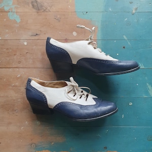 Könnte beinhalten: Ein Paar zweifarbige Schuhe im Vintage-Stil. Die Schuhe haben ein weißes Obermaterial mit marineblauen Details an Zehen, Ferse und Seiten. Sie haben ein Schnürdesign und einen kleinen, schwarzen Absatz. Die Schuhe sind vor einem Holzgrund platziert.