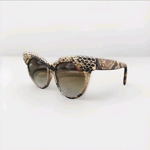Peut inclure: Lunettes de soleil œil de chat avec un motif peau de serpent dans les tons marron, beige et noir. Les lunettes ont des verres teintés et un design élégant et vintage. La monture et les branches sont entièrement recouvertes du motif peau de serpent.