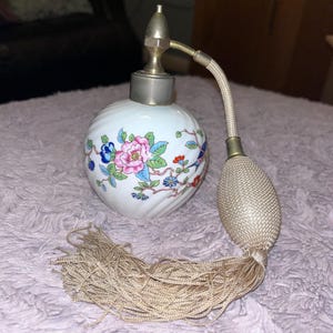 May include: A vintage perfume atomizer with a white ceramic body decorated with floral patterns in pink, blue, and green. It features a silver-toned metal top and a beige bulb with a matching tassel.