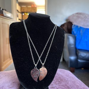 May include: Two heart-shaped pendant necklaces displayed on a black bust. Each pendant is a different color, one silver and one copper, with a floral design. The necklaces have silver chains.