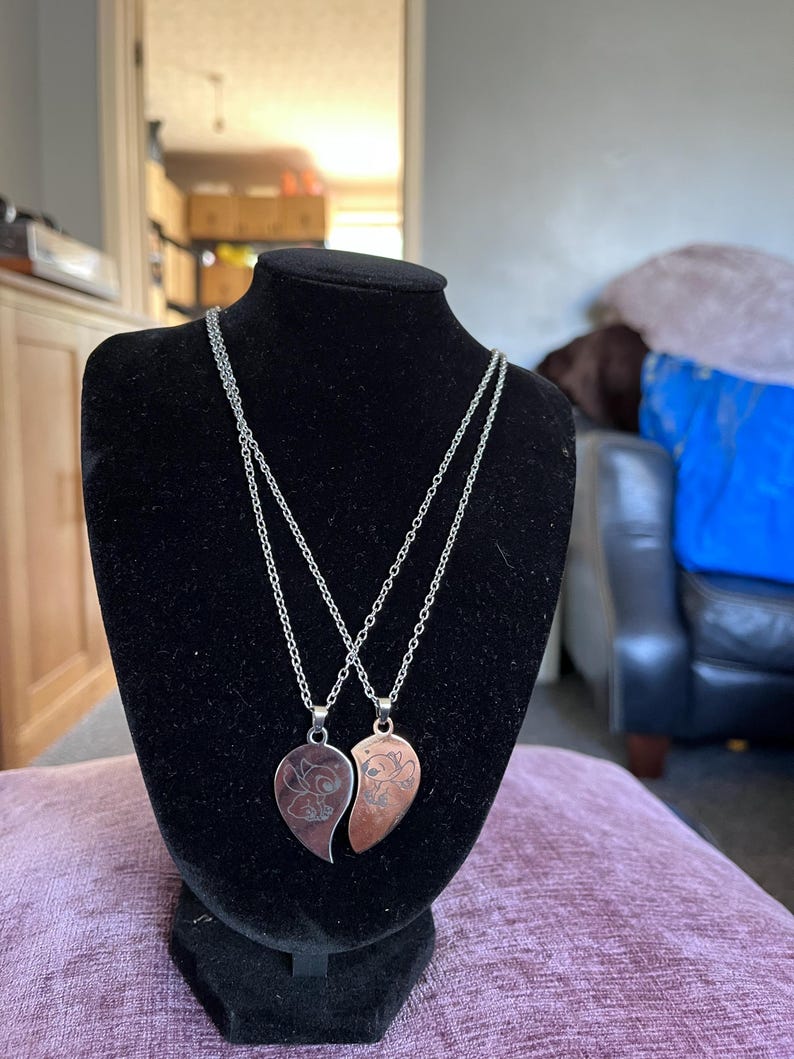 May include: Two heart-shaped pendant necklaces displayed on a black bust. Each pendant is a different color, one silver and one copper, with a floral design. The necklaces have silver chains.