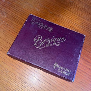 May include: A vintage deck of Waddington's Bezique playing cards in a purple box. The box has gold lettering that reads "Waddington's Bezique" and "Playing Cards". The box is rectangular and appears to be made of cardboard.