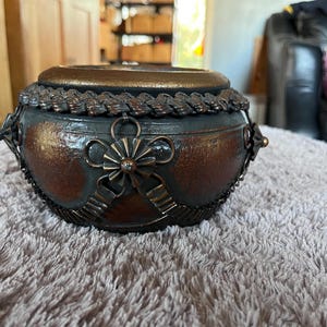 May include: A decorative, round container with a lid. The container is primarily bronze and dark gray, with ornate metalwork around the rim and sides. A bow-shaped embellishment with a central jewel adorns the front. The container sits on a light gray, textured surface.
