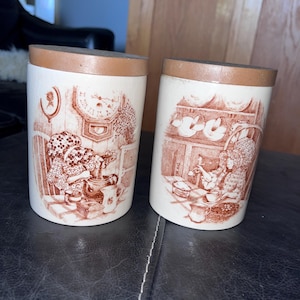 May include: Two ceramic canisters with brown lids and sepia-toned illustrations. One depicts a girl with a clock, the other a girl in a kitchen. The canisters are white with brown accents.