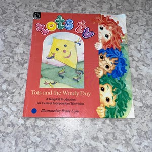 Könnte beinhalten: Ein Kinderbuch mit dem Titel "Tots and the Windy Day" mit rotem Einband. Das Cover zeigt einen gelben Drachen und Illustrationen von drei Zeichentrickfiguren mit bunten Haaren.