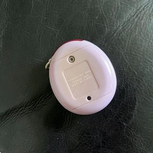 May include: A light pink, egg-shaped electronic pet device with a metal keychain loop on the side. The back features a battery compartment cover secured by a single screw, with text reading BANDAI 2004 MADE IN CHINA embossed on the plastic surface.