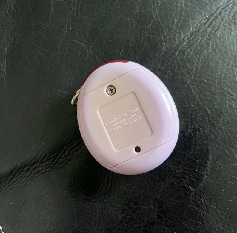 May include: A light pink, egg-shaped electronic pet device with a metal keychain loop on the side. The back features a battery compartment cover secured by a single screw, with text reading BANDAI 2004 MADE IN CHINA embossed on the plastic surface.