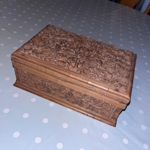May include: A rectangular wooden box with detailed floral carvings on the lid and sides. The box is a warm brown colour, possibly hardwood. The carvings show leaves and flowers, indicating a decorative storage item, possibly for jewellery.