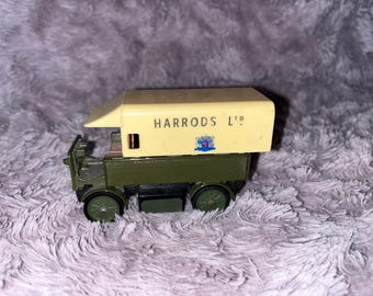 Puede incluir: Un camión de juguete vintage con una parte superior color crema y una base verde oliva. La parte superior está impresa con "HARRODS L½o" y un escudo. El camión tiene ruedas negras. Un juguete de colección.