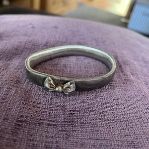 Puede incluir: Un brazalete plateado con un lazo decorativo. El brazalete tiene una textura acanalada y un pequeño lazo en el centro. La joya se muestra sobre una superficie morada.