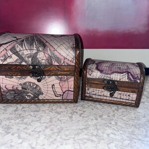 May include: Two arched-top storage chests featuring a vintage map print in shades of pink, cream, and black. Each box has dark brown wooden trim with a rope-like texture, a metal latch closure, and visible cartographic details on the surface.