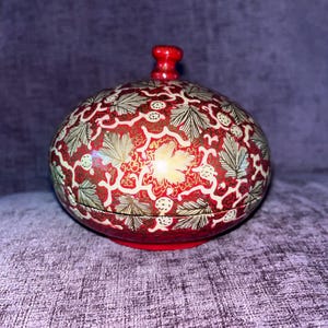 Pode incluir: Uma caixa decorativa redonda vermelha com tampa. A caixa é adornada com um padrão detalhado de folhas douradas e brancas e desenhos florais sobre um fundo vermelho. A tampa tem um pequeno botão vermelho.