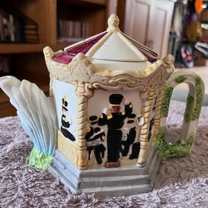 May include: A decorative teapot with a carousel design. The teapot features a white body with a maroon and white roof, and a handle with a green vine design. The teapot has a detailed illustration of a marching band.