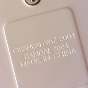 May include: A light pink plastic surface featuring a recessed rectangular area with embossed text that reads BANDAI WIZ 2004, BANDAI 2004, and MADE IN CHINA. A single silver-colored metal screw is visible at the top center of the pink plastic casing.