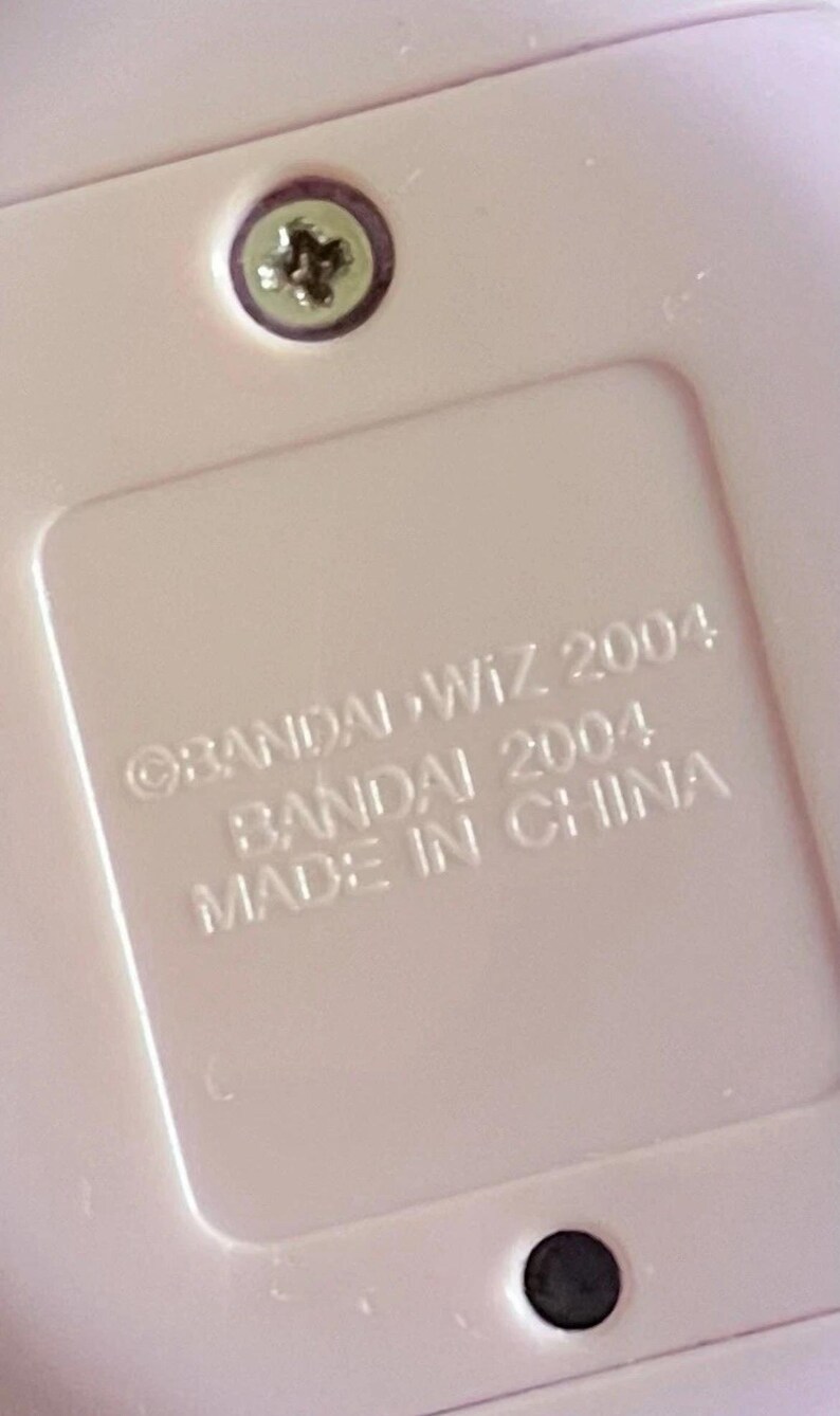 May include: A light pink plastic surface featuring a recessed rectangular area with embossed text that reads BANDAI WIZ 2004, BANDAI 2004, and MADE IN CHINA. A single silver-colored metal screw is visible at the top center of the pink plastic casing.