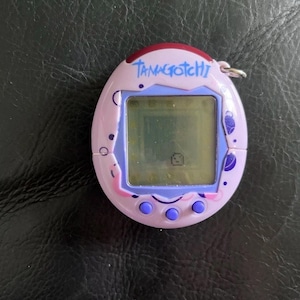May include: A light purple Tamagotchi digital pet device with a square screen displaying a small pixelated character. The oval-shaped plastic shell features dark purple circular patterns, a dark red top accent, and three round purple buttons below.