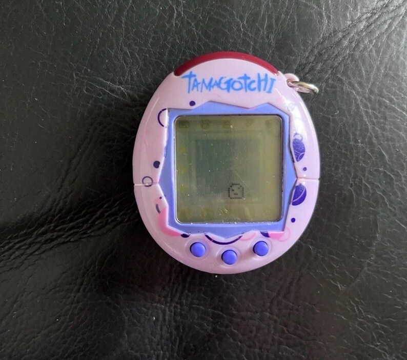 May include: A light purple Tamagotchi digital pet device with a square screen displaying a small pixelated character. The oval-shaped plastic shell features dark purple circular patterns, a dark red top accent, and three round purple buttons below.