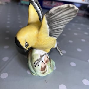 以下が含まれることがあります： 黒とグレーの翼を持つ黄色の鳥の陶器製の置物。緑と茶色の台座に止まっています。鳥はダイナミックなポーズで、翼は部分的に広がっています。置物は細かく、手描きのように見えます。