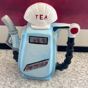 May include: A novelty teapot shaped like a gas pump, painted light blue with black accents. The teapot has a white top with the word "TEA" in red. The words "GAS STATION" are also visible. The digital display reads "14.7".