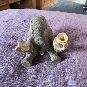 May include: A grey elephant ornament with a textured finish, holding two small decorative pots. The pots have a beige base and colourful accents. The elephant is sitting on a purple textured surface.