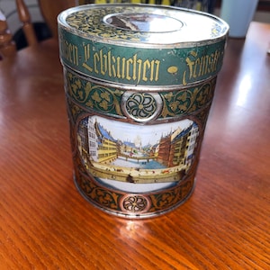 May include: A vintage tin with a green and gold design. The tin features a detailed illustration of a European city scene. The lid and body are adorned with ornate patterns and text. The tin is cylindrical and sits on a wooden surface.