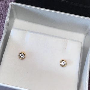 May include: A pair of gold stud earrings with a clear, round gemstone set in each. The earrings are displayed in a white box with a silver trim, resting on a purple surface. The box contains the text "Tiphereth, 8 Butcher Row, Beverley, Tel:- 01482886186".