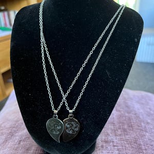 May include: Two silver-colored necklaces with heart-shaped pendants, each featuring a cartoon character design. The pendants are split to form a complete heart when joined. The necklaces are displayed on a black mannequin.