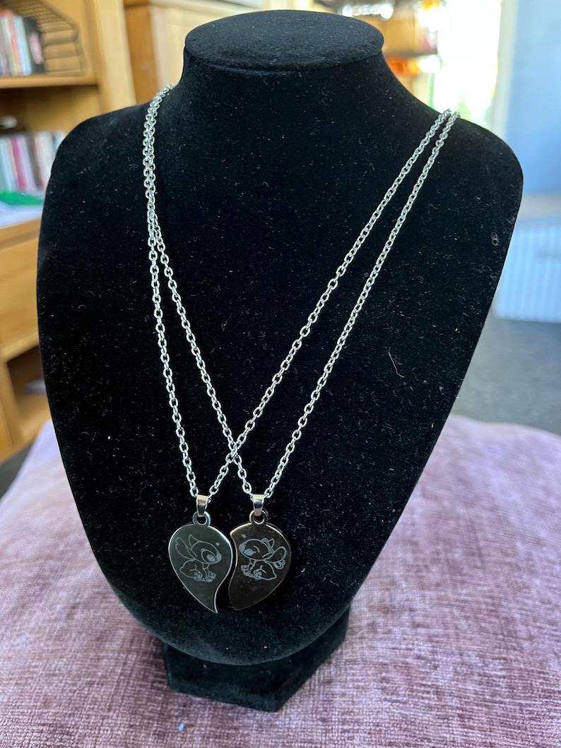 May include: Two silver-colored necklaces with heart-shaped pendants, each featuring a cartoon character design. The pendants are split to form a complete heart when joined. The necklaces are displayed on a black mannequin.