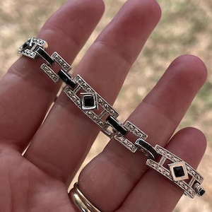 May include: Silver bracelet with a geometric design. The bracelet has alternating rectangular and square links, with black enamel and small clear stones. The bracelet is held in a hand.