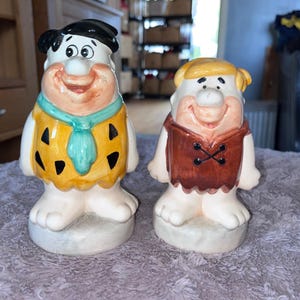 Könnte beinhalten: Zwei Keramikfiguren von Zeichentrickfiguren, eine mit schwarzen Haaren und einem gelb-schwarzen Outfit, die andere mit blonden Haaren und einer braunen Weste. Beide haben lächelnde Gesichter und stehen auf einem hellfarbigen Sockel.