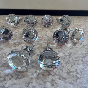 Puede incluir: Una colección de pomos de cristal transparente y facetado para armarios. Los pomos tienen un aspecto de diamante, reflejando la luz. El herraje está diseñado para proyectos de mobiliario y decoración del hogar. Los pomos están dispuestos sobre una superficie moteada.