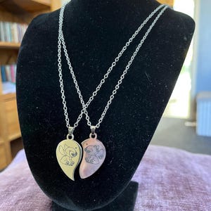 May include: Two-piece heart pendant necklaces with a cartoon character design. Each pendant is a half-heart shape, one gold-toned and the other copper-toned, with a silver chain. The necklaces are displayed on a black velvet bust.