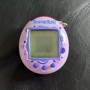 May include: A light purple Tamagotchi digital pet device with a screen displaying a small character. The oval-shaped plastic shell features dark purple circular patterns and the word TAMAGOTCHI printed in blue at the top. Three purple buttons are below.