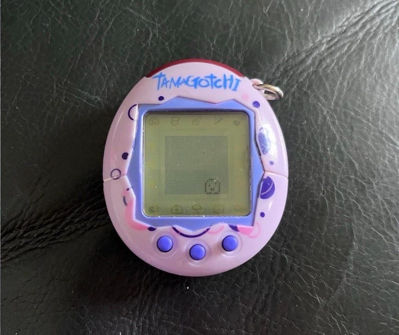 May include: A light purple Tamagotchi digital pet device with a screen displaying a small character. The oval-shaped plastic shell features dark purple circular patterns and the word TAMAGOTCHI printed in blue at the top. Three purple buttons are below.
