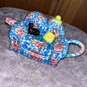Könnte beinhalten: Eine skurrile Teekanne in Form eines Sofas, verziert mit einem blauen, roten und weißen Spritzmuster. Eine kleine schwarze Katzenfigur sitzt auf dem Sofa, zusammen mit einem gelben Kissen und Miniaturbüchern. Die Teekanne hat einen gelben Hut.