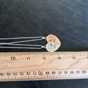 May include: Two heart-shaped pendant necklaces. Each pendant is split into two halves, with a cartoon character design on each half. The necklaces have silver chains. A wooden ruler is placed next to the necklaces, with measurements in inches.