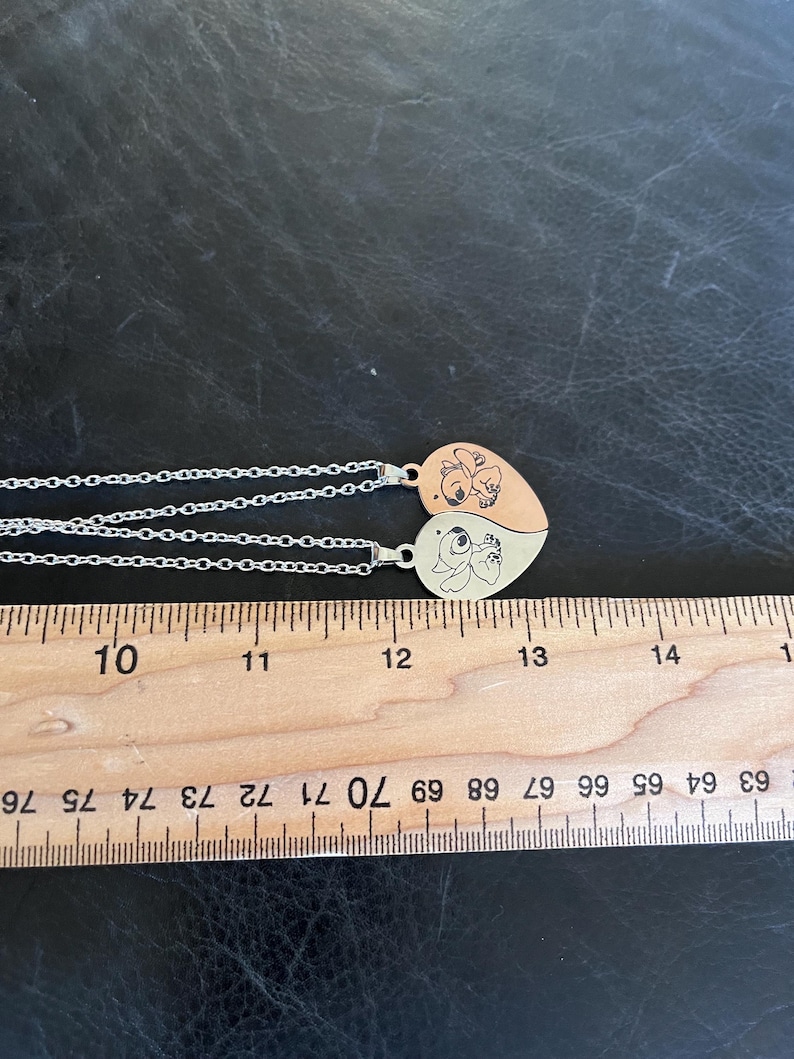 May include: Two heart-shaped pendant necklaces. Each pendant is split into two halves, with a cartoon character design on each half. The necklaces have silver chains. A wooden ruler is placed next to the necklaces, with measurements in inches.