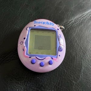 May include: A light purple Tamagotchi digital pet device with a square LCD screen, three round purple buttons below the display, and a metal keychain ring attached to the top. The casing features dark purple circular and swirl graphic patterns.