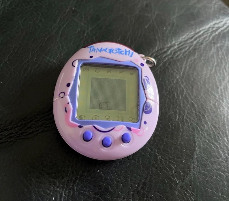 May include: A light purple Tamagotchi digital pet device with a square LCD screen, three round purple buttons below the display, and a metal keychain ring attached to the top. The casing features dark purple circular and swirl graphic patterns.