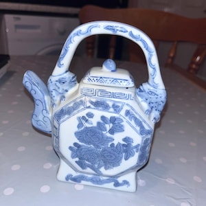 Può includere: Una piccola teiera in ceramica bianca e blu con un motivo floreale. La teiera ha un corpo ottagonale, un manico curvo e un piccolo beccuccio. Il motivo floreale è blu e copre il corpo della teiera. Il manico e il beccuccio hanno un motivo blu e bianco.