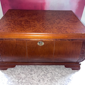 May include: A rectangular wooden chest with a rich brown finish and a burl wood veneer top. The front features a central brass-toned keyhole escutcheon and carved decorative side panels. The chest stands on four short, sturdy feet.